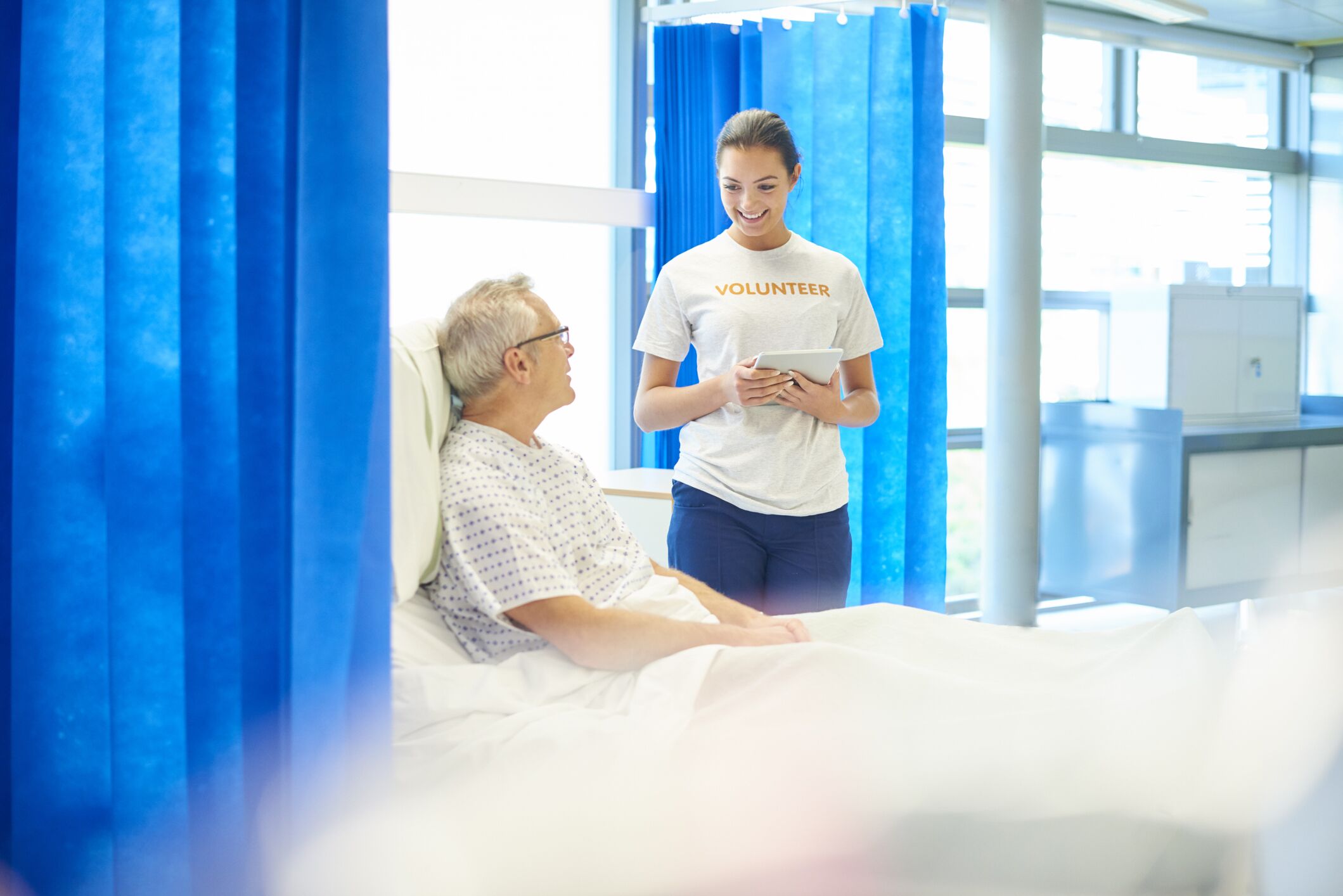 Alt text: Hospital volunteer with a tablet smiling and talking to an elderly patient in a hospital bed, surrounded by blue curtains and natural light.