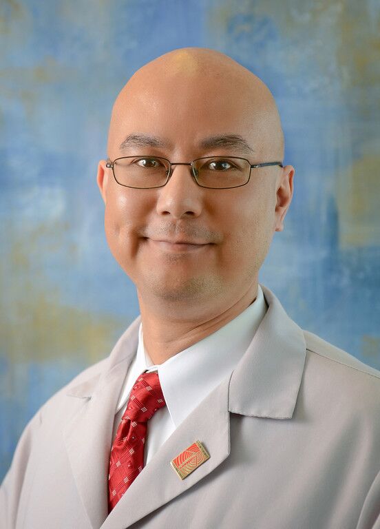 A person with glasses and a bald head wearing a white lab coat and a red tie stands against a blue and yellow textured background.