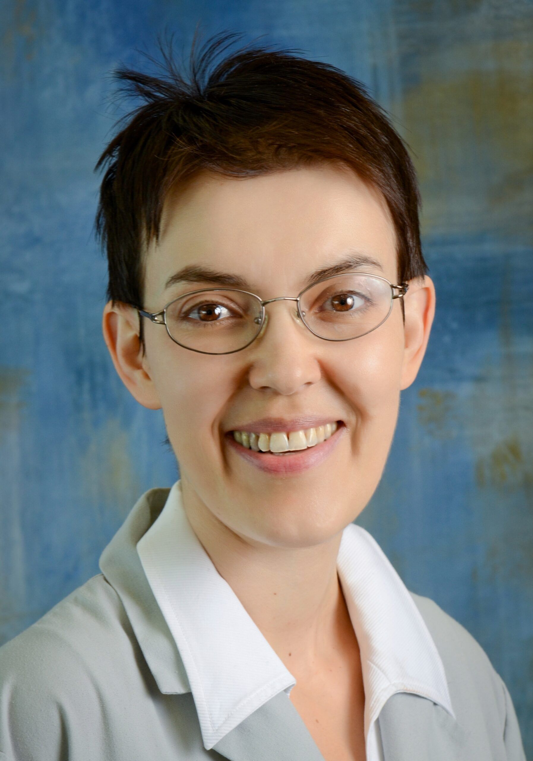 Person with short dark hair and glasses smiling, wearing a white-collared shirt and gray jacket. The background is a mottled blue and brown.