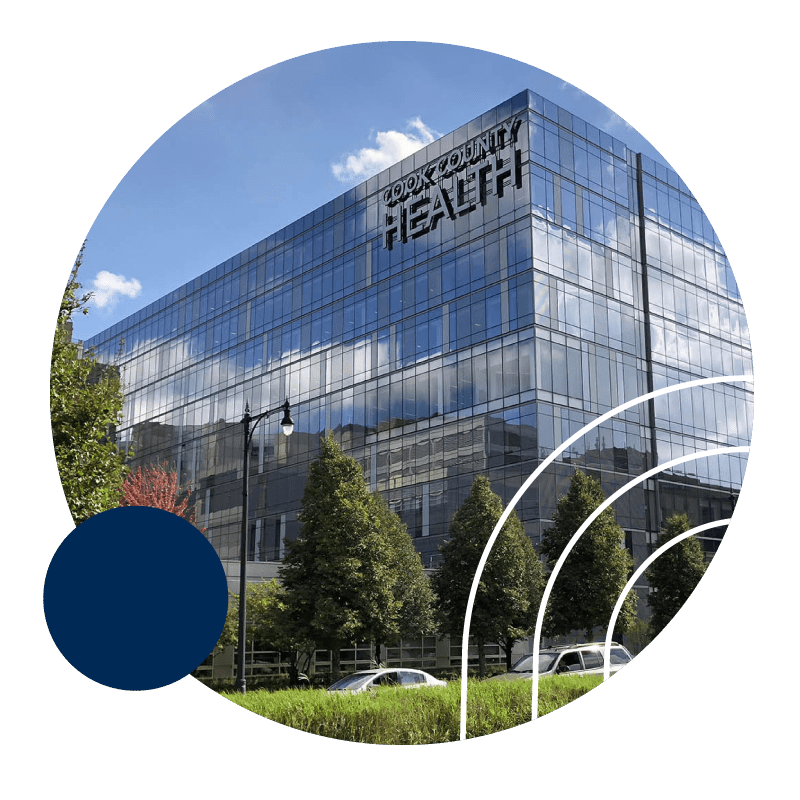A modern glass building marked Cook County Health is shown against a clear blue sky. The facade reflects clouds and is surrounded by trees and a curved structure in the foreground.