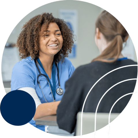 A smiling healthcare professional in blue scrubs, wearing a stethoscope, sits at a table with a patient who has long brown hair. They appear to be engaged in a conversation in a clinical setting.