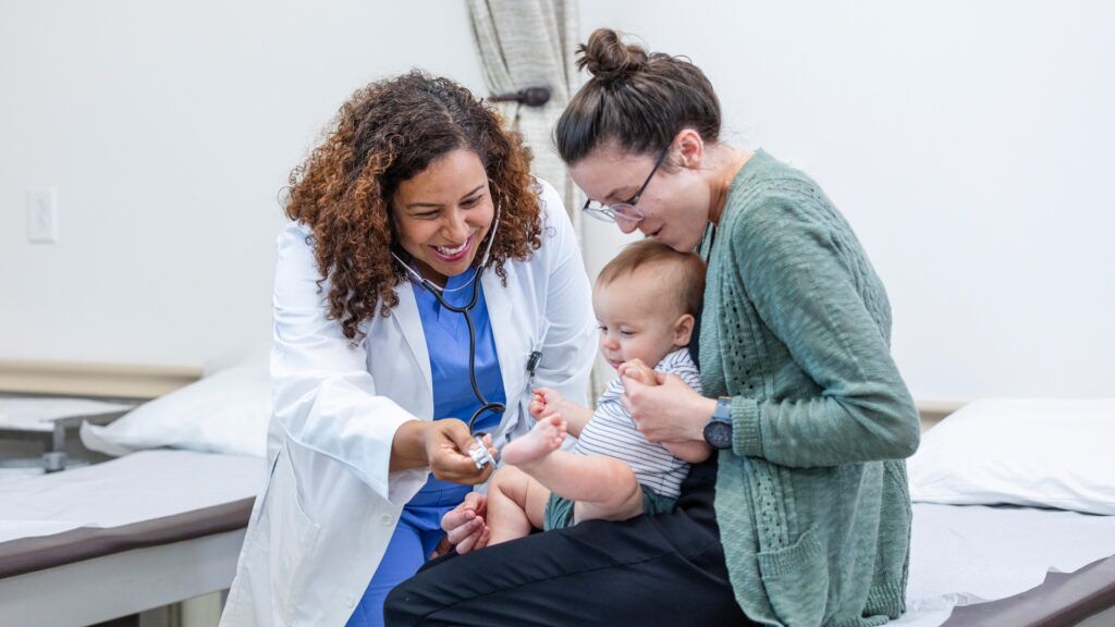 A doctor in a white coat and blue scrubs smiles while examining a baby held by a woman. The woman, wearing a green cardigan, sits on an examination table. The baby is grabbing a medical instrument with interest.
