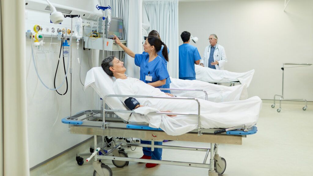 A hospital room with two patients lying in beds. A nurse in blue scrubs checks the monitors, while a doctor in a white coat stands near the second patient. The room is well-lit and equipped with medical devices.